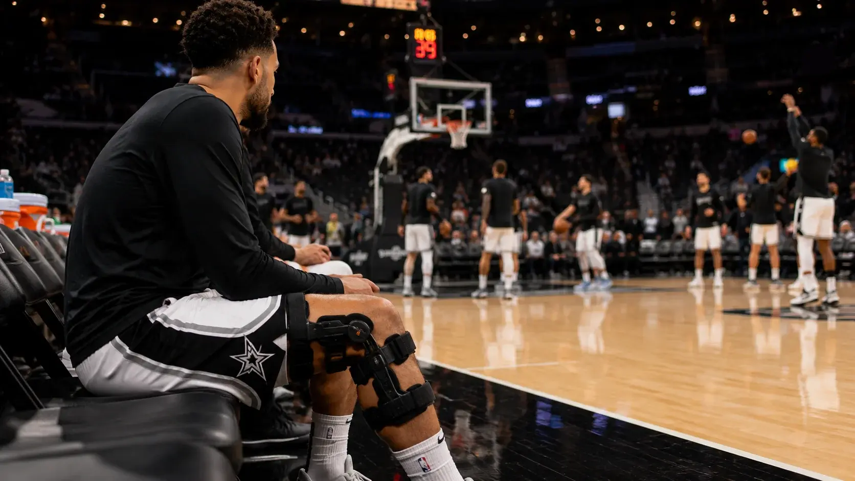 NBA player on the bench with a knee brace watching teammates warm up before a game