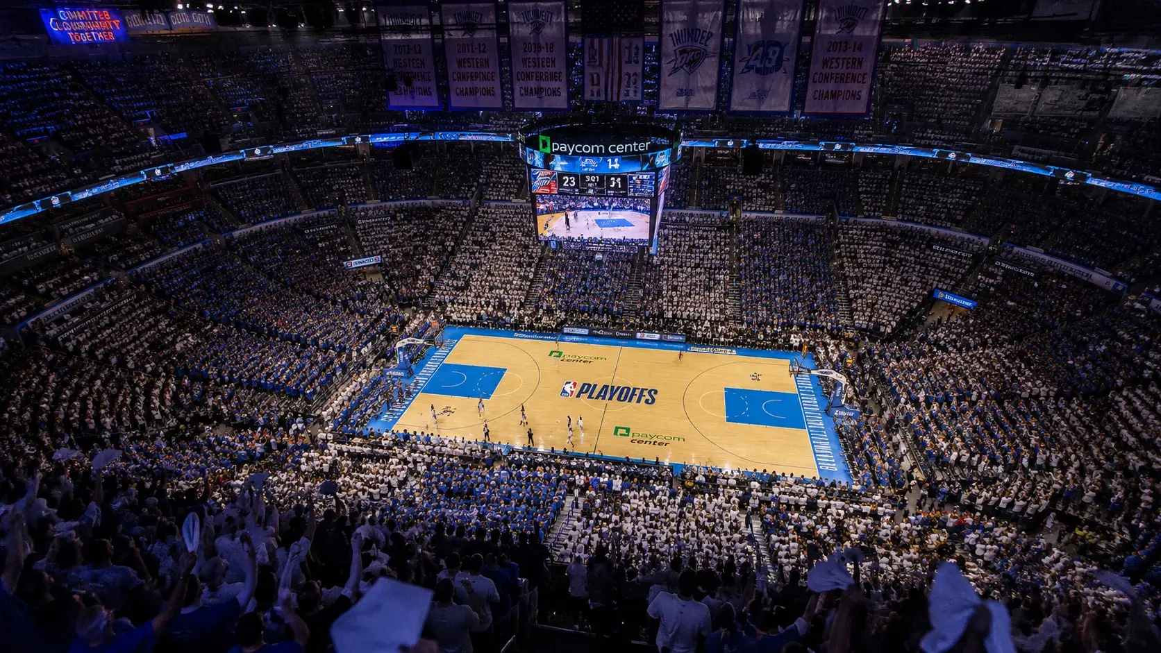 NBA playoff bracket displayed on a large arena screen with a packed crowd and championship banners overhead